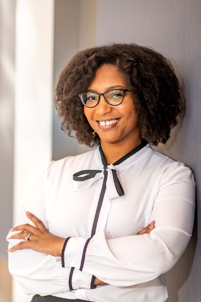 An African-American presenting woman is smiling. She is wearing a white blouse and glasses and has natural hair. 