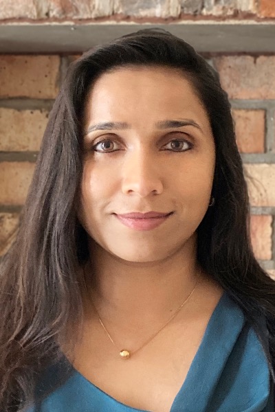 A brown woman is smiling. She has dark hair and is wearing a blue blouse. A necklace is showing.