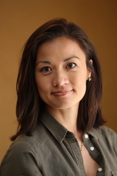 An Asian-presenting woman is smiling. She is wearing a greenish brown dress shirt with the top three buttons all open. The background is a mixture of brown and yellow.