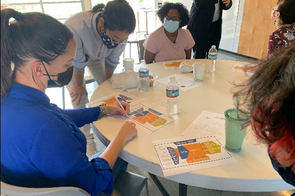 Student sit/stand around a rounded white table; a staff makes notes on printout paper;