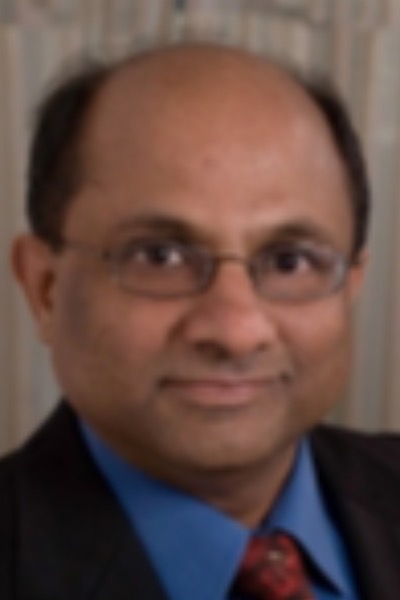 A headshot of a brown-skinned man wearing glasses with no hair on top and little hair on the sides. He is smiling and wearing a dark suit jacket with a blue shirt and red tie.