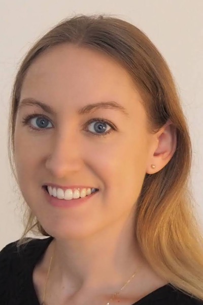 A headshot of a white woman. She has long brown hair and is smiling. She is wearing a black blouse and a gold necklace.