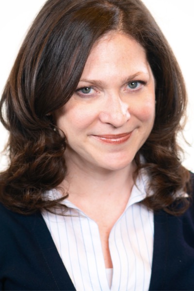 A headshot of a white woman. She has long brown hair and is smiling. She is wearing a white blouse and a black jacket.