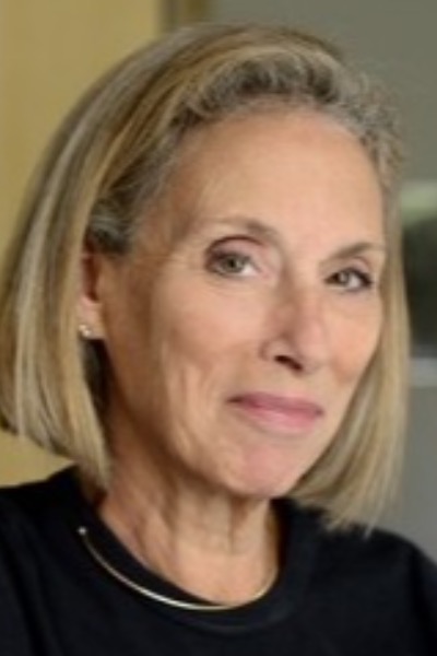 A headshot of a white woman. She has medium dirty blond hair and is smiling. She is wearing a black blouse and a necklace.