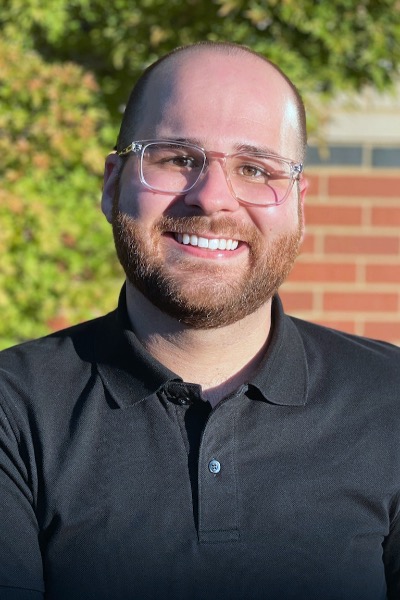Headshot of a smiling white male with very short brown hair and a thin beard and mustache. He is wearing eyeglasses and a black polo shirt.
