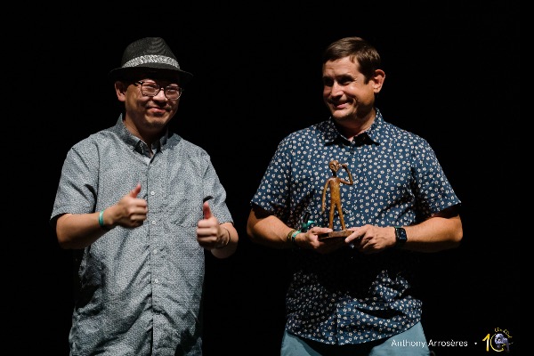 Two men are shown from the waist up. Left: Asian-presenting, fedora and glasses, making the double thumbs up sign. Right: white-presenting, smiling, and holding up a statuette. Background is solid black. Photo credit at bottom right: Anthony Arroseres