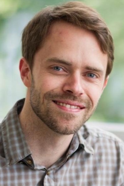 A caucasian-presenting man is smiling. He has short brown hair and a thin brown beard. He is wearing a plaid brown dress shirt.